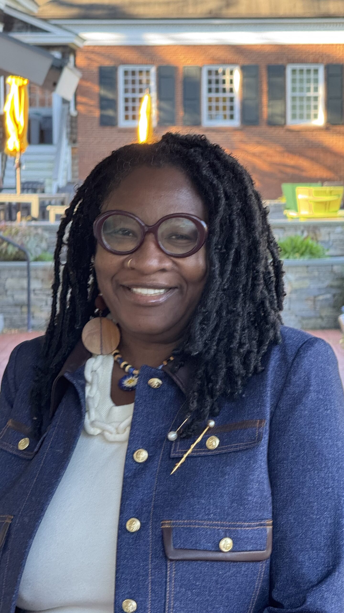 A smiling woman with dark locs wearing round glasses and a dark blue jacket.
