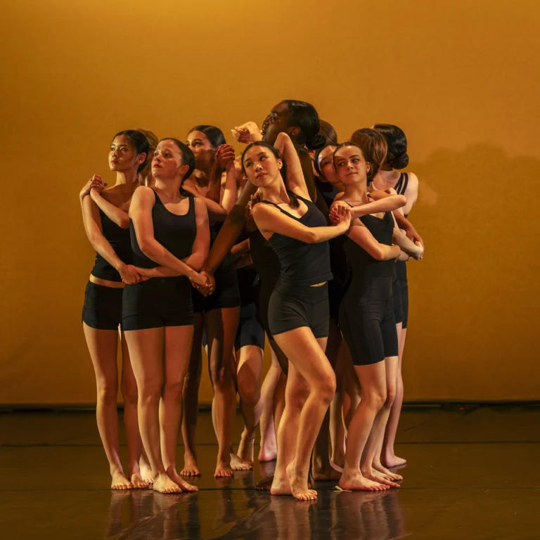 A group of dancers in black leotards and shorts stand in a tight, interconnected cluster against an orange background.