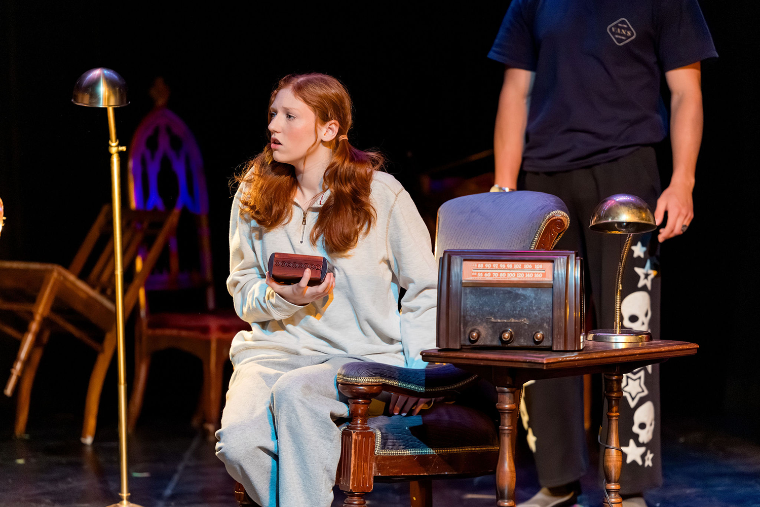 A young woman with long auburn hair sits in a chair on a stage, holding a small box, while an antique radio sits on a side table next to her.