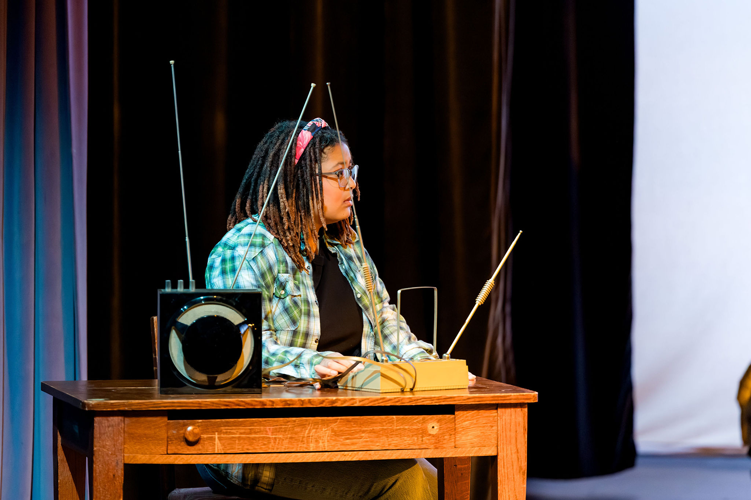 A person with dreadlocks and glasses sits at a desk, playing a theremin equipped with multiple metal antennas.