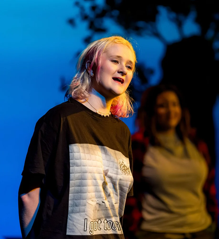 A young person with blonde hair singing on a stage, with two people blurred in the background.