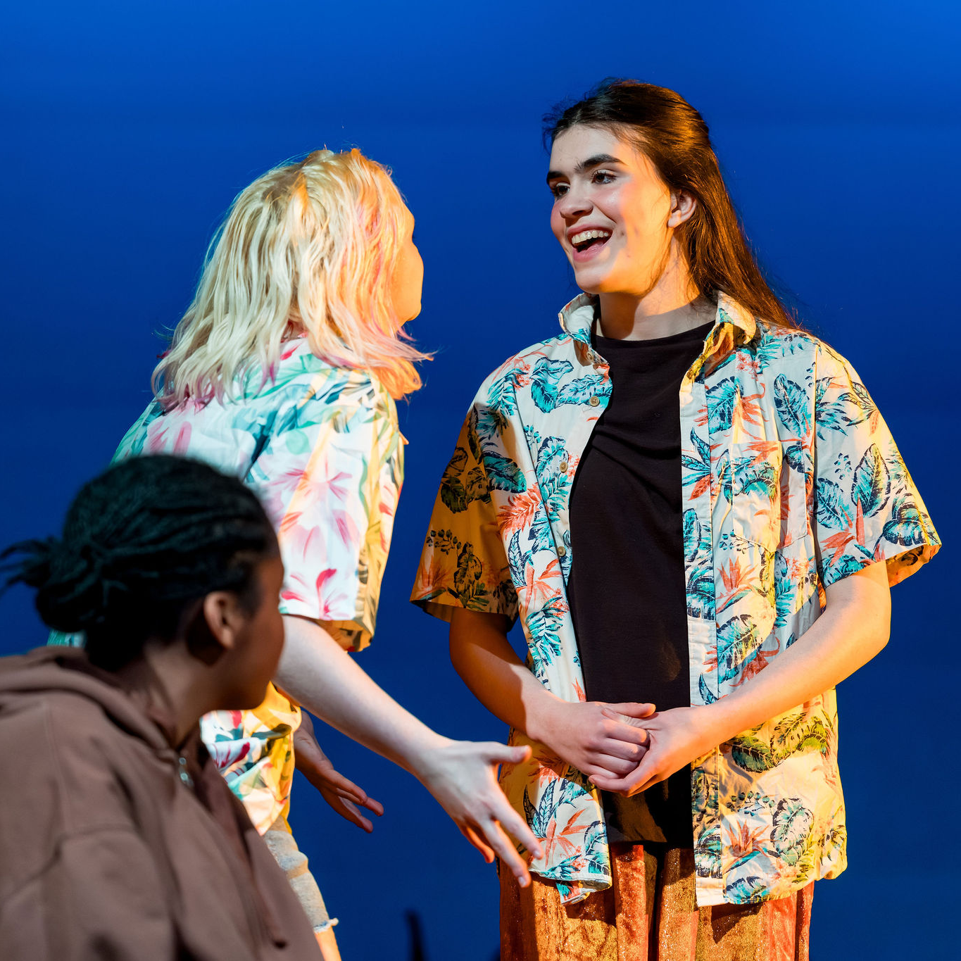 Three actors on stage in a theatrical performance; one standing actor wearing a tropical print shirt laughs while speaking to another, with a third actor seated in the foreground.