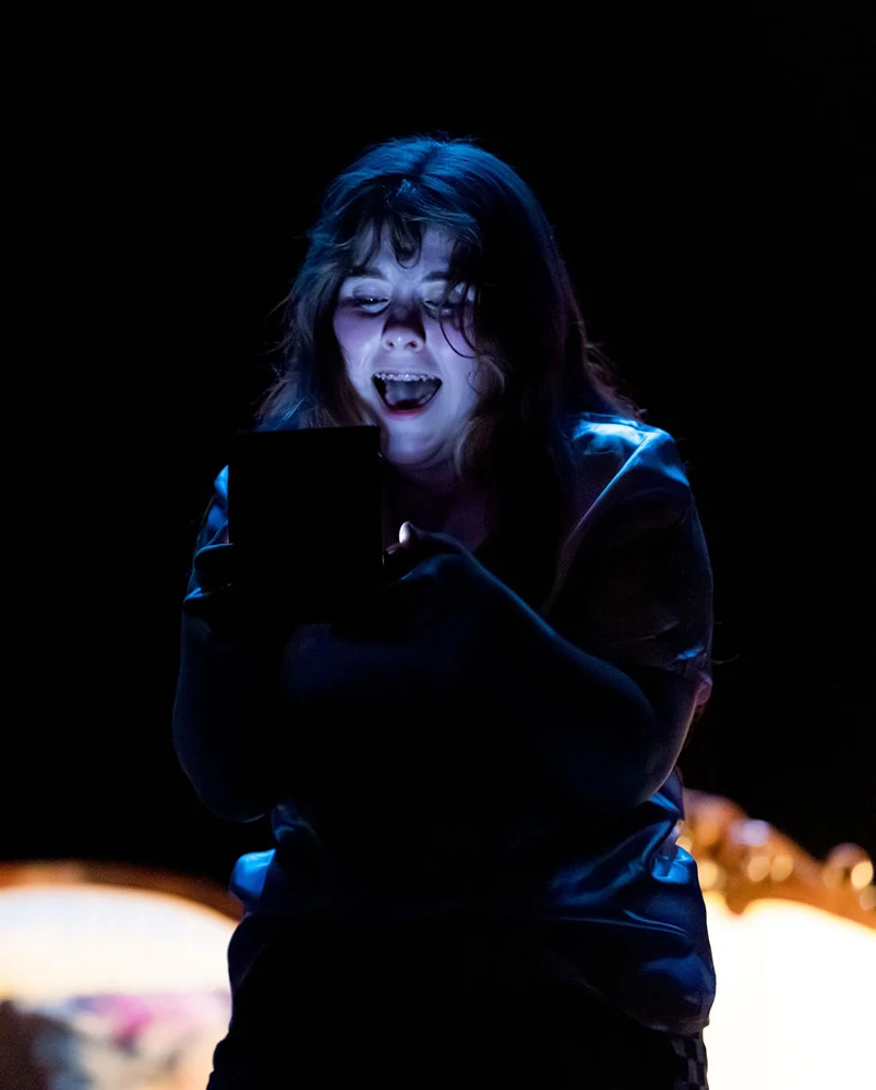 A young person with a surprised expression is illuminated by the glow of a smartphone in a dark room.