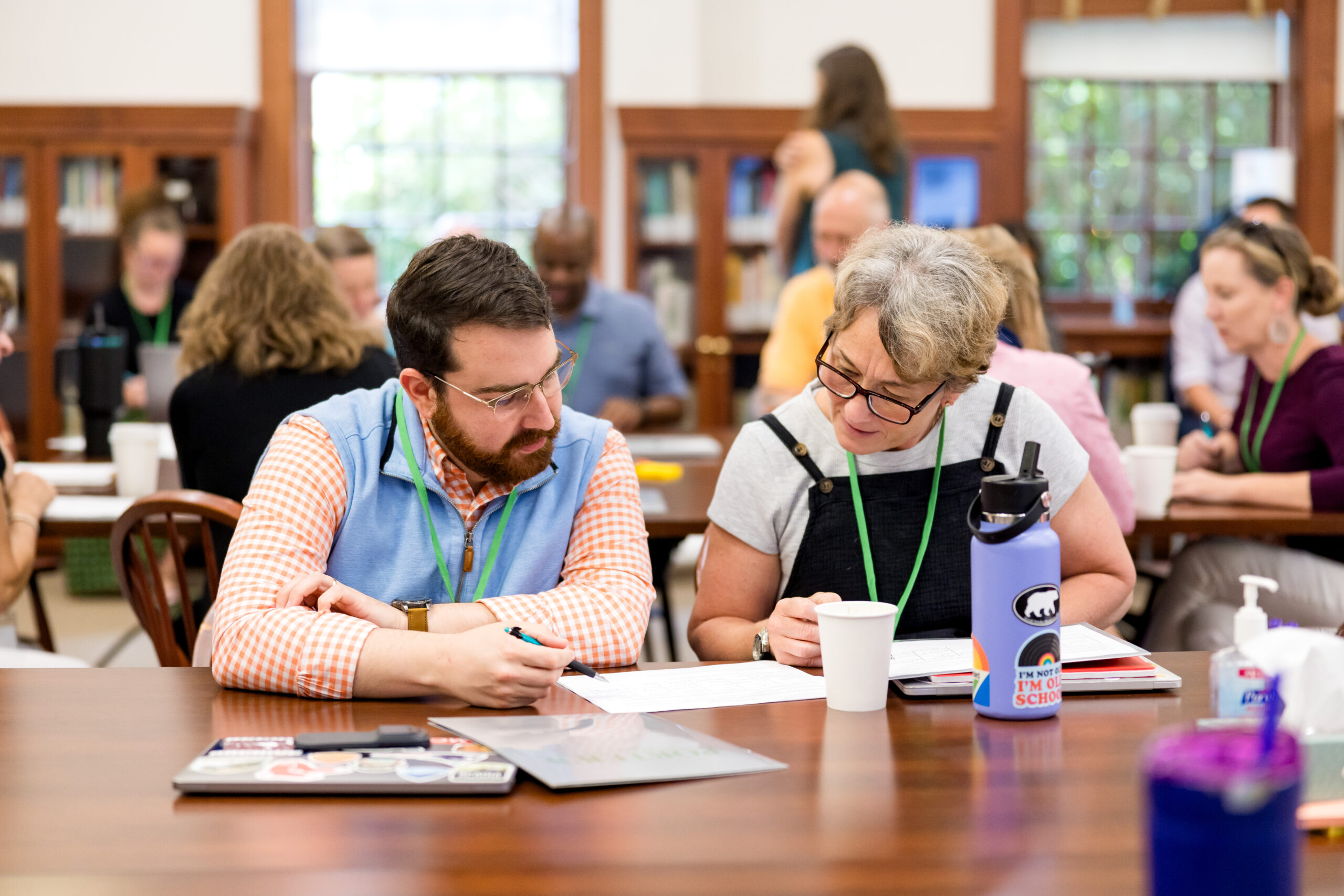 A man and woman sit at a wooden table in a library setting, looking down at a document while working together.