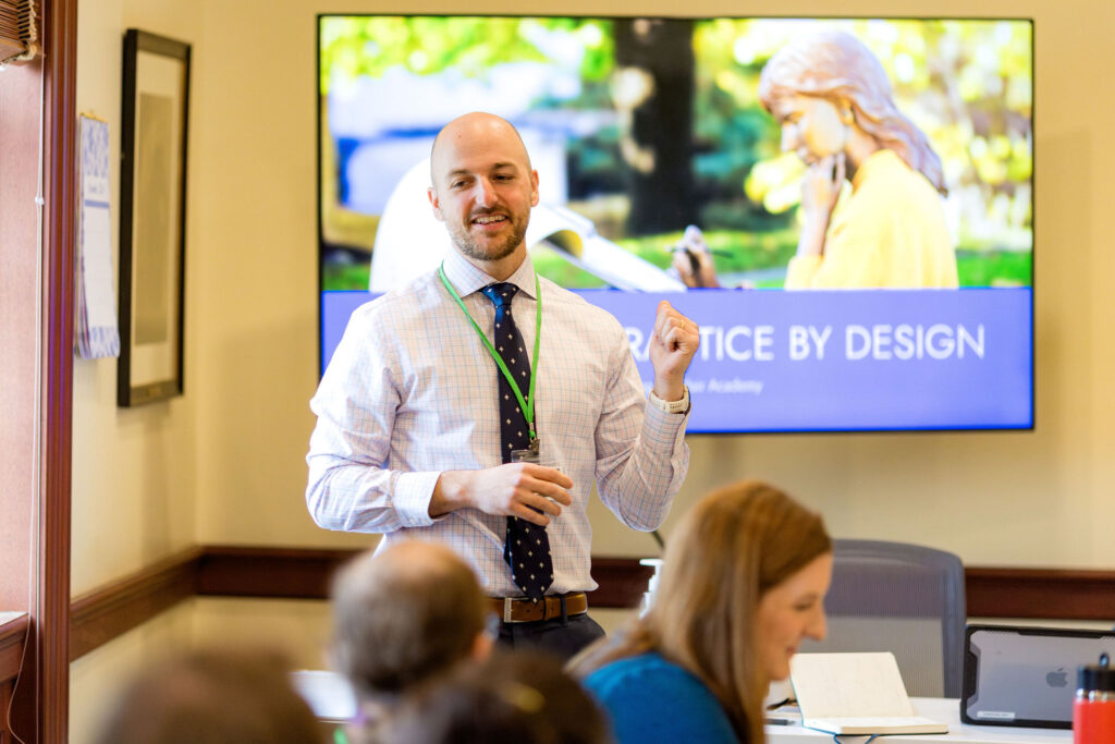 A man in a dress shirt and tie gestures while presenting to a group; a screen behind him reads PRACTICE BY DESIGN with a photo of a woman outdoors.