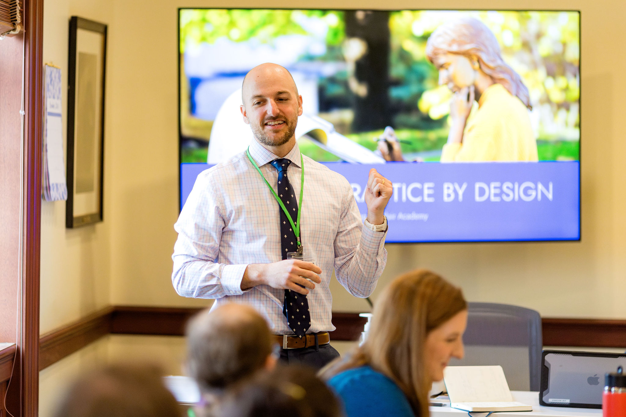 A man in a dress shirt and tie gestures while presenting to a group; a screen behind him reads PRACTICE BY DESIGN with a photo of a woman outdoors.