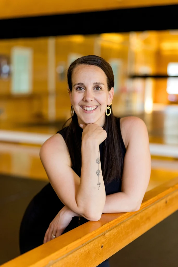 A smiling woman with a tattoo on her forearm rests her chin on her hand while leaning on a wooden ballet barre in a dance studio.