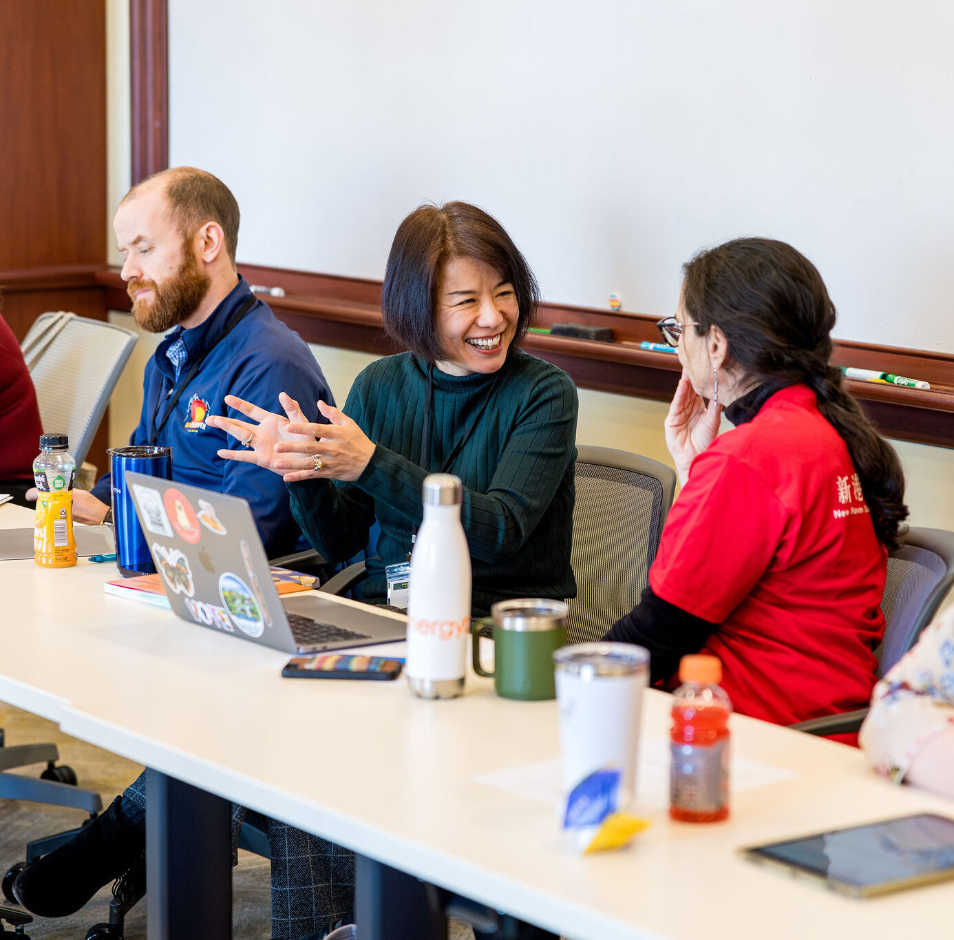 People sit at a long table in a meeting room, talking and smiling. Laptops, drinks, and papers are on the table. One woman gestures enthusiastically while others listen and engage in conversation.