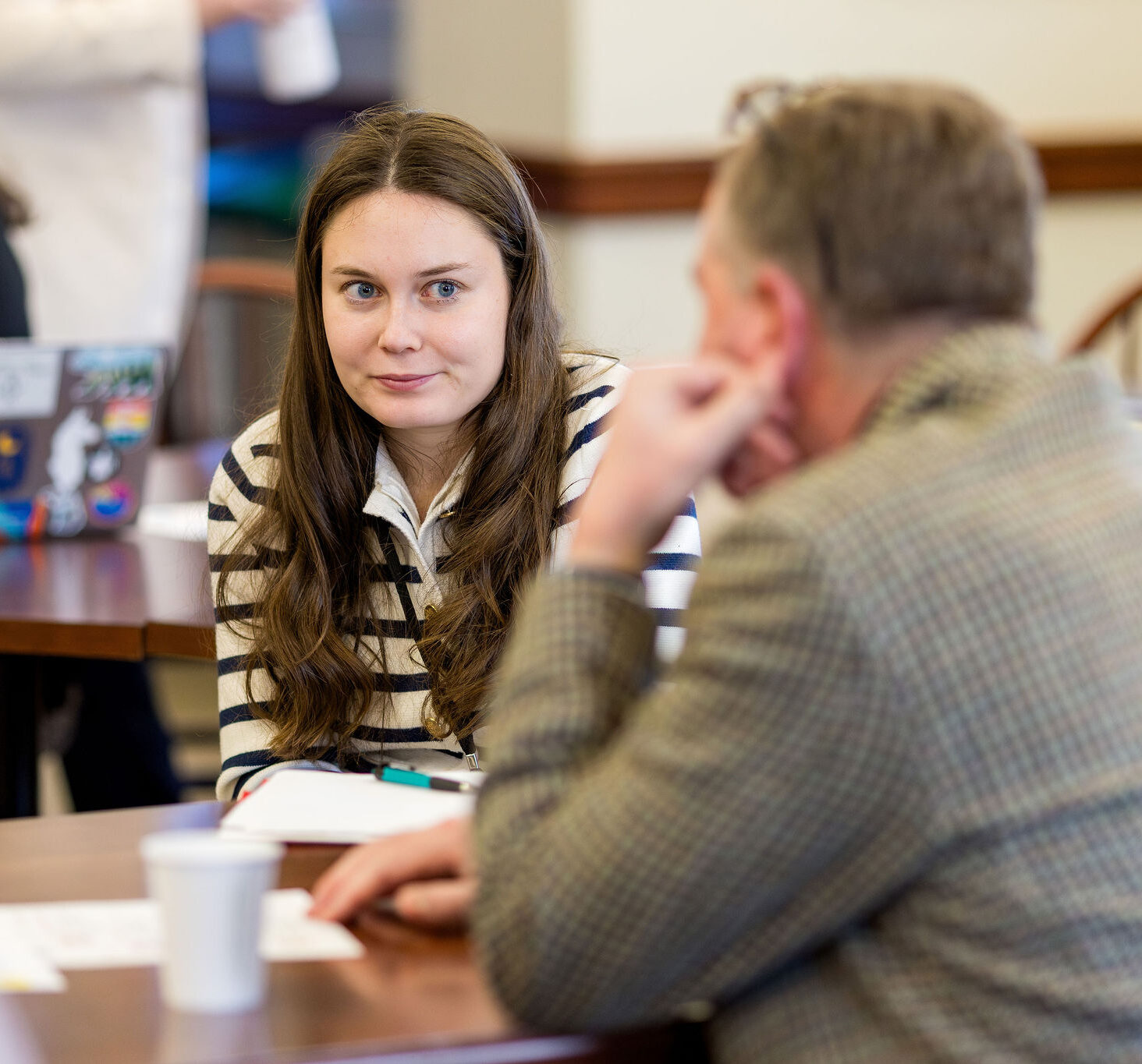 A woman with long brown hair in a striped shirt listens attentively to a man in a blazer across a table in a classroom or meeting setting. Papers and coffee cups are on the table. Other people are in the background.