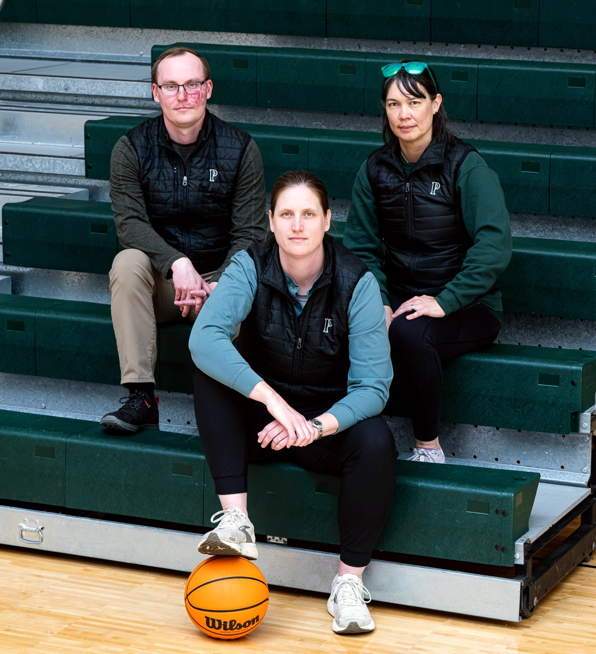 Three adults pose on gym bleachers, wearing dark vests and long-sleeved shirts, with a basketball on the floor in front of them. The bleachers and floor are green and wood-colored.