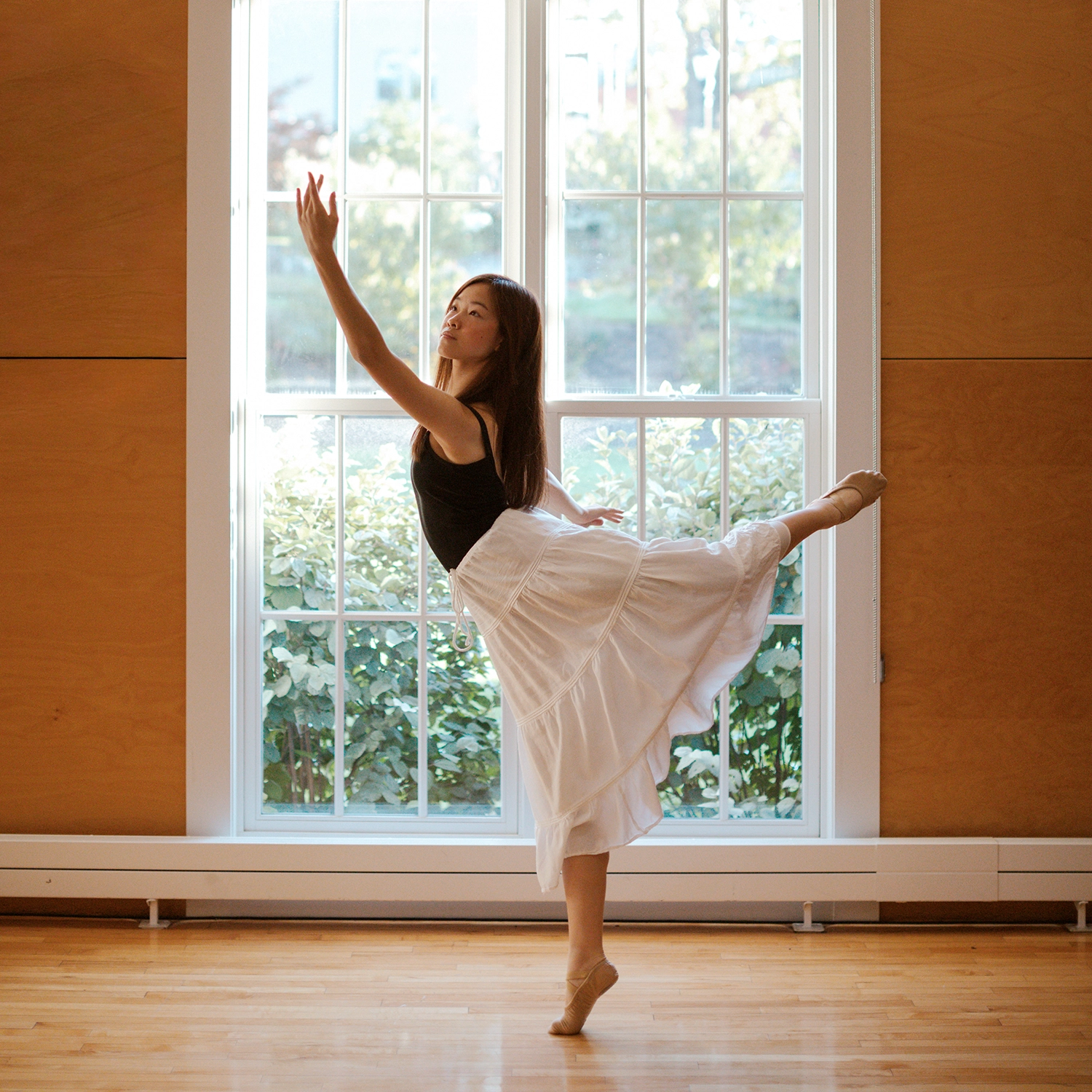 A ballet dancer in a black tank top and white skirt poses in an arabesque in front of a large window.
